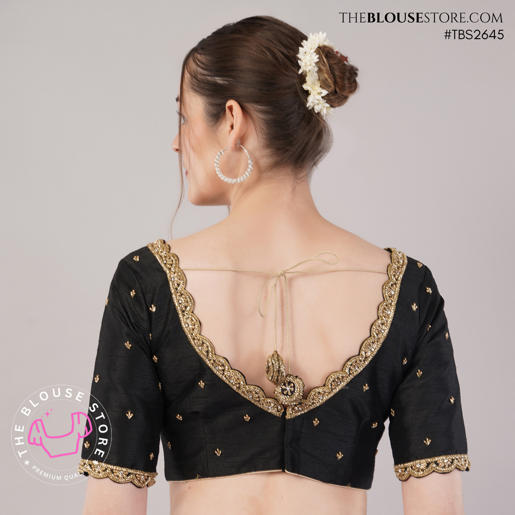 A woman shown from the back wearing a black short-sleeve blouse with gold embroidered trim, a deep open back, and tie details. The photo is taken against a light gray studio background with visible TheBlouseStore branding and watermark.