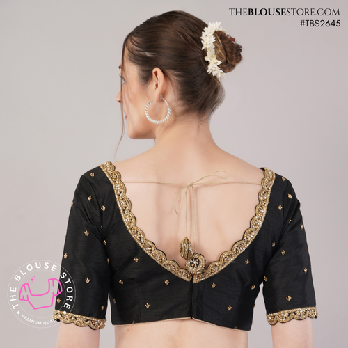 A woman shown from the back wearing a black short-sleeve blouse with gold embroidered trim, a deep open back, and tie details. The photo is taken against a light gray studio background with visible TheBlouseStore branding and watermark.