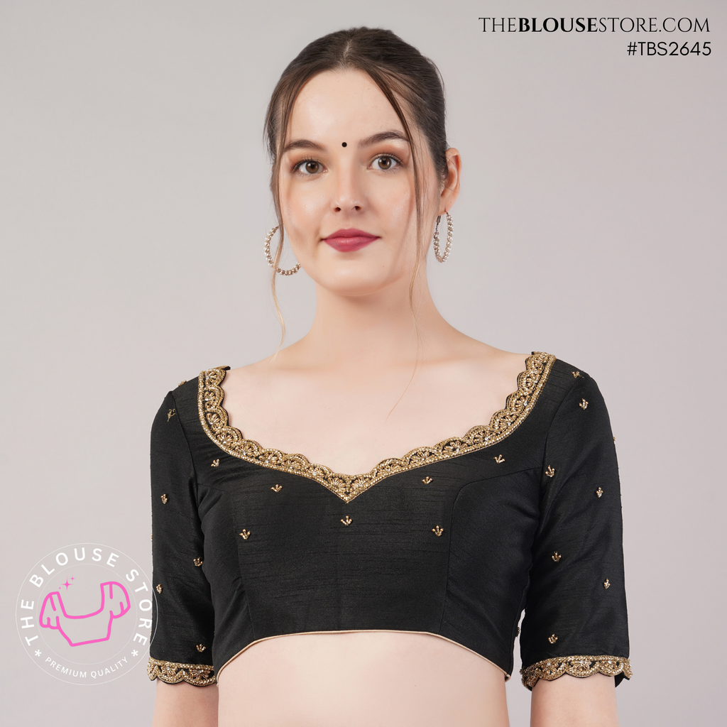A woman modeling a black short-sleeve blouse with gold embroidered border detailing along the scoop neckline and sleeve cuffs, set against a plain light gray studio background.