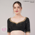 A woman modeling a black short-sleeve blouse with gold embroidered border detailing along the scoop neckline and sleeve cuffs, set against a plain light gray studio background.