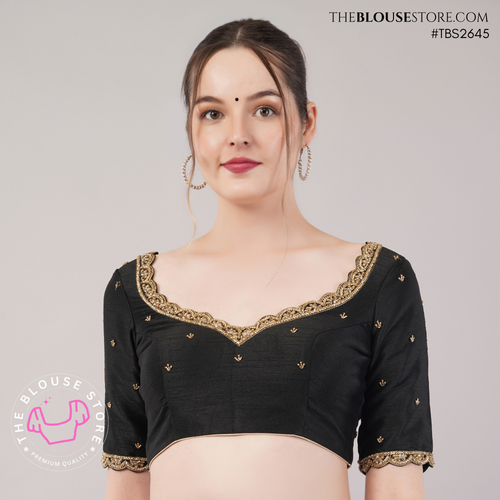 A woman modeling a black short-sleeve blouse with gold embroidered border detailing along the scoop neckline and sleeve cuffs, set against a plain light gray studio background.
